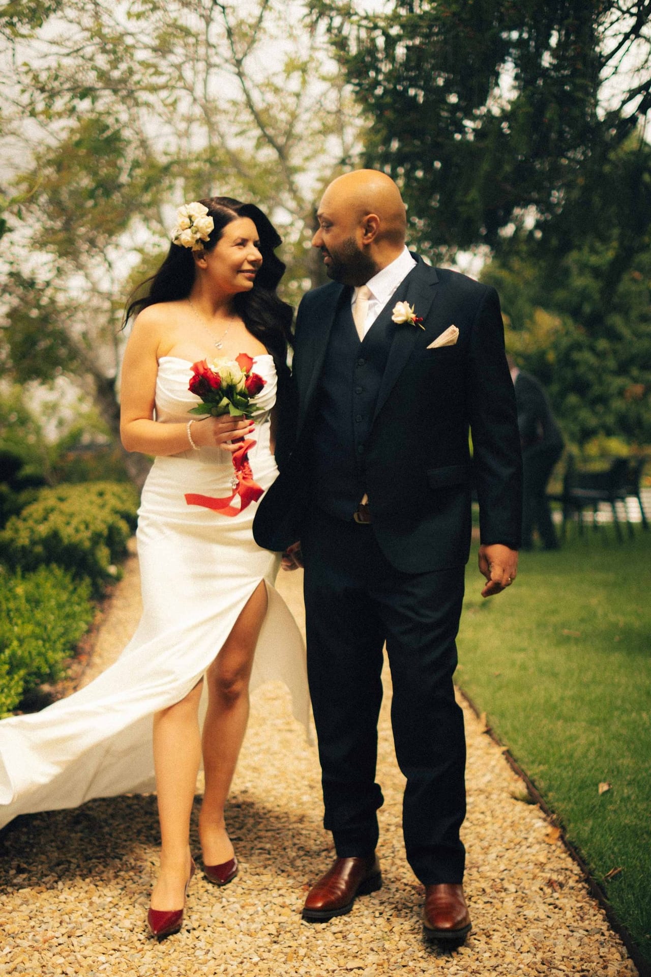 A couple walking hand in hand along a garden path, the bride holding red roses