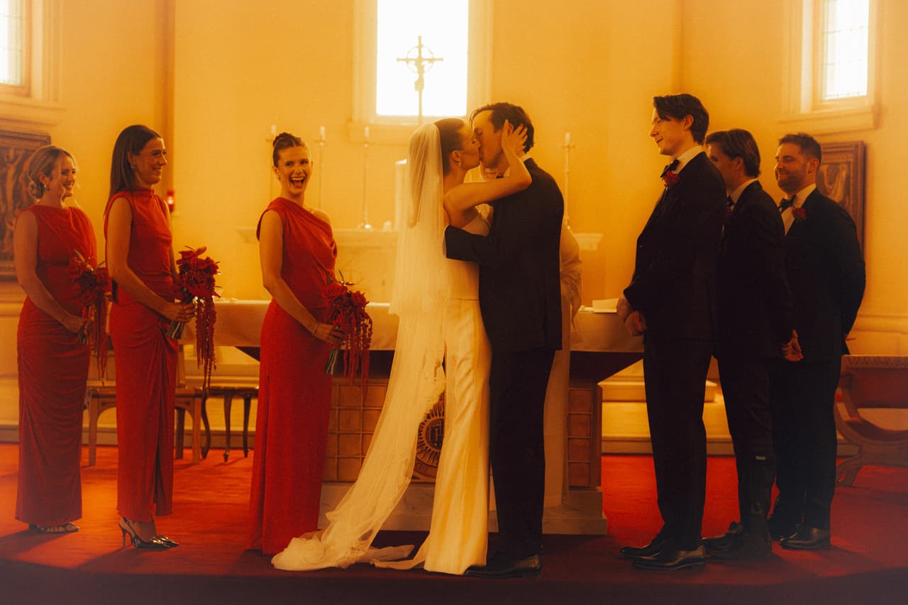 A couple kissing at the altar of a sunlit church, bridesmaids in red on one side and groomsmen on the other