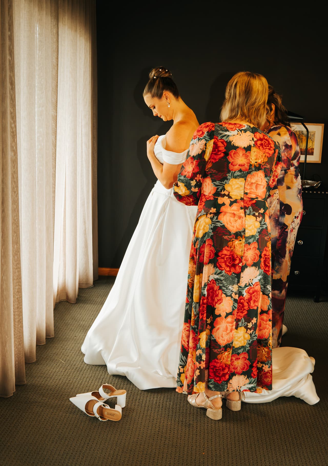 A bride getting ready while a woman in a floral dress adjusts her gown