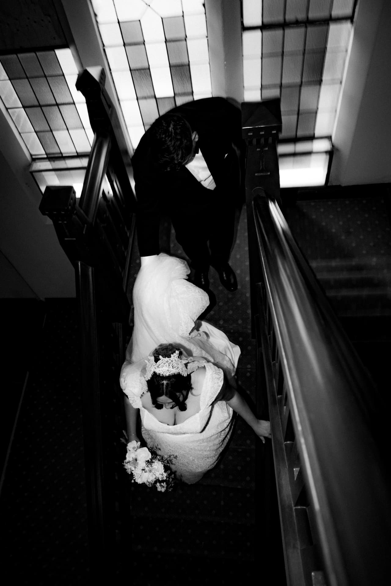 Black and white photo of a couple seen from above on a staircase, her veil and bouquet in the light
