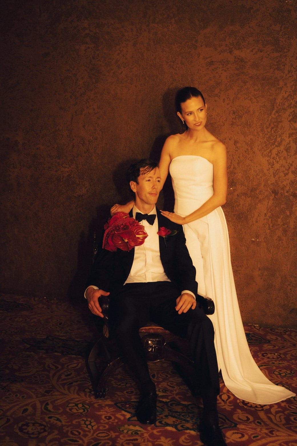 A bride standing beside the seated groom, who holds red roses, in an ornate theatre room