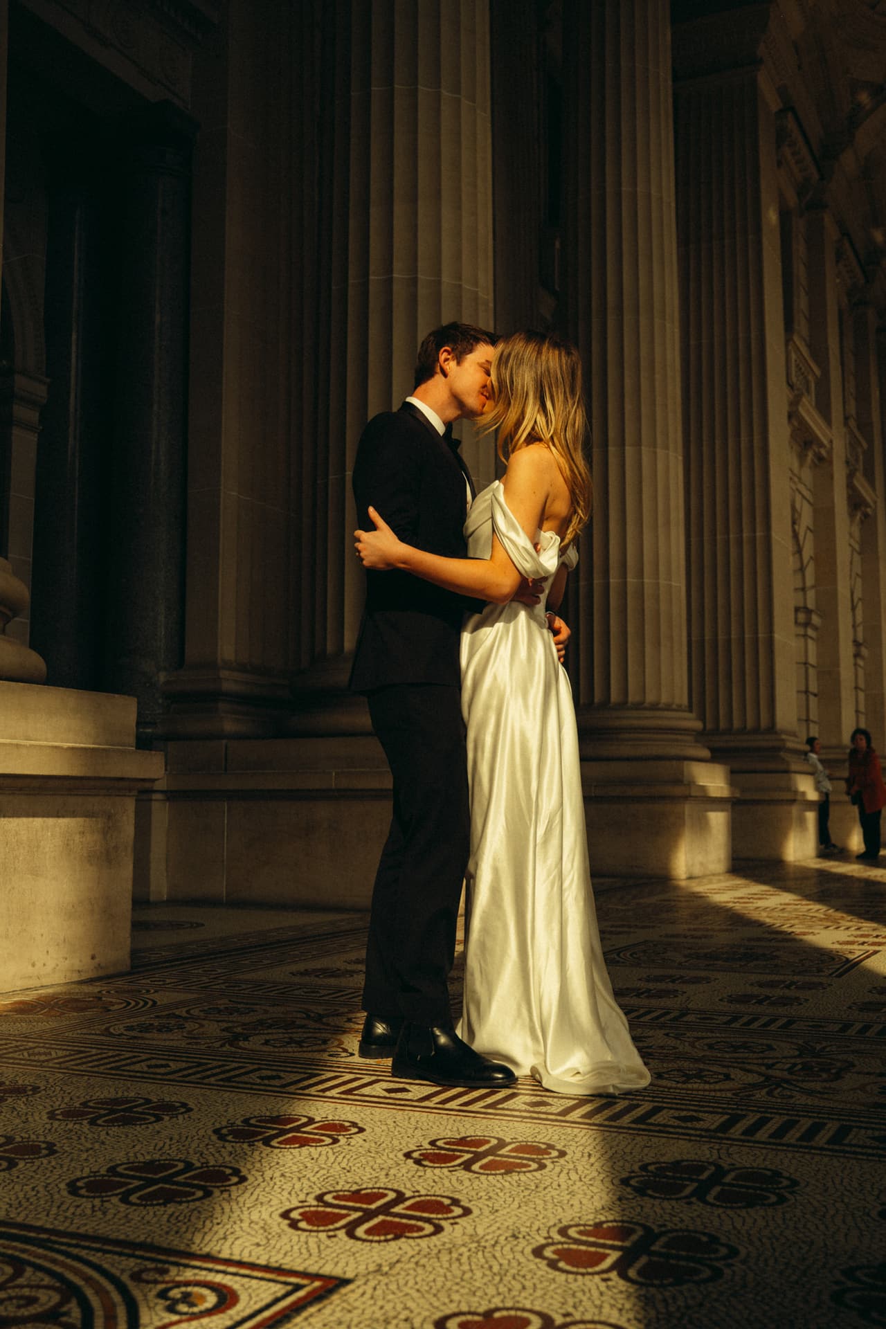 Hannah and Josh kissing in the colonnade, sunlight across the patterned tiles