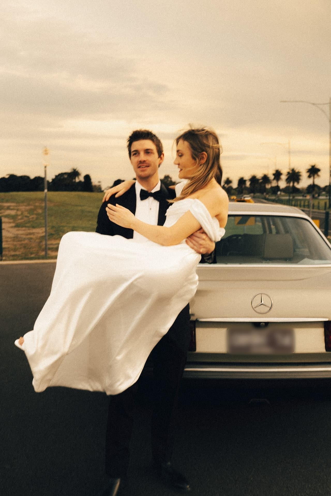 Josh lifting Hannah in his arms beside the vintage Mercedes, both laughing