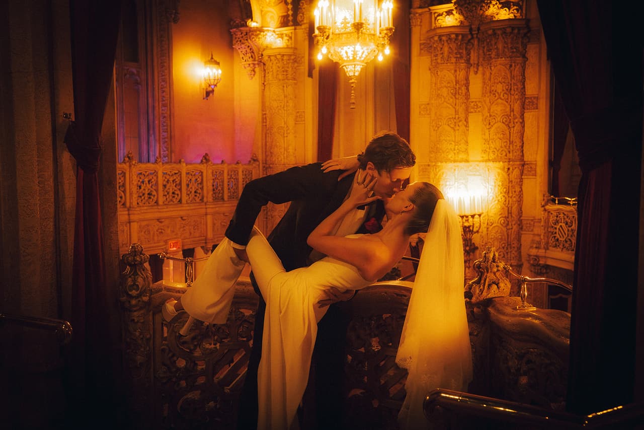 Nick leaning in to kiss Kira in a candlelit corner of the Regent Theatre, a chandelier glowing behind them